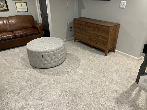 Carpet installation in basement with a brown leather couch, grey ottoman, and wood-look dresser.
